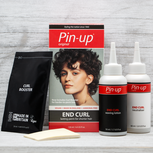 An End Curl Perm kit by Pin-up Perm on a white wooden surface. The kit includes a box featuring a woman with curly hair, a bottle of waving lotion, a bottle of neutralizer, a sachet of curl booster, and a sponge. Labeled "Vegan" and "Made in Britain," this set delivers the perfect permanent curling for your Pin-up style.